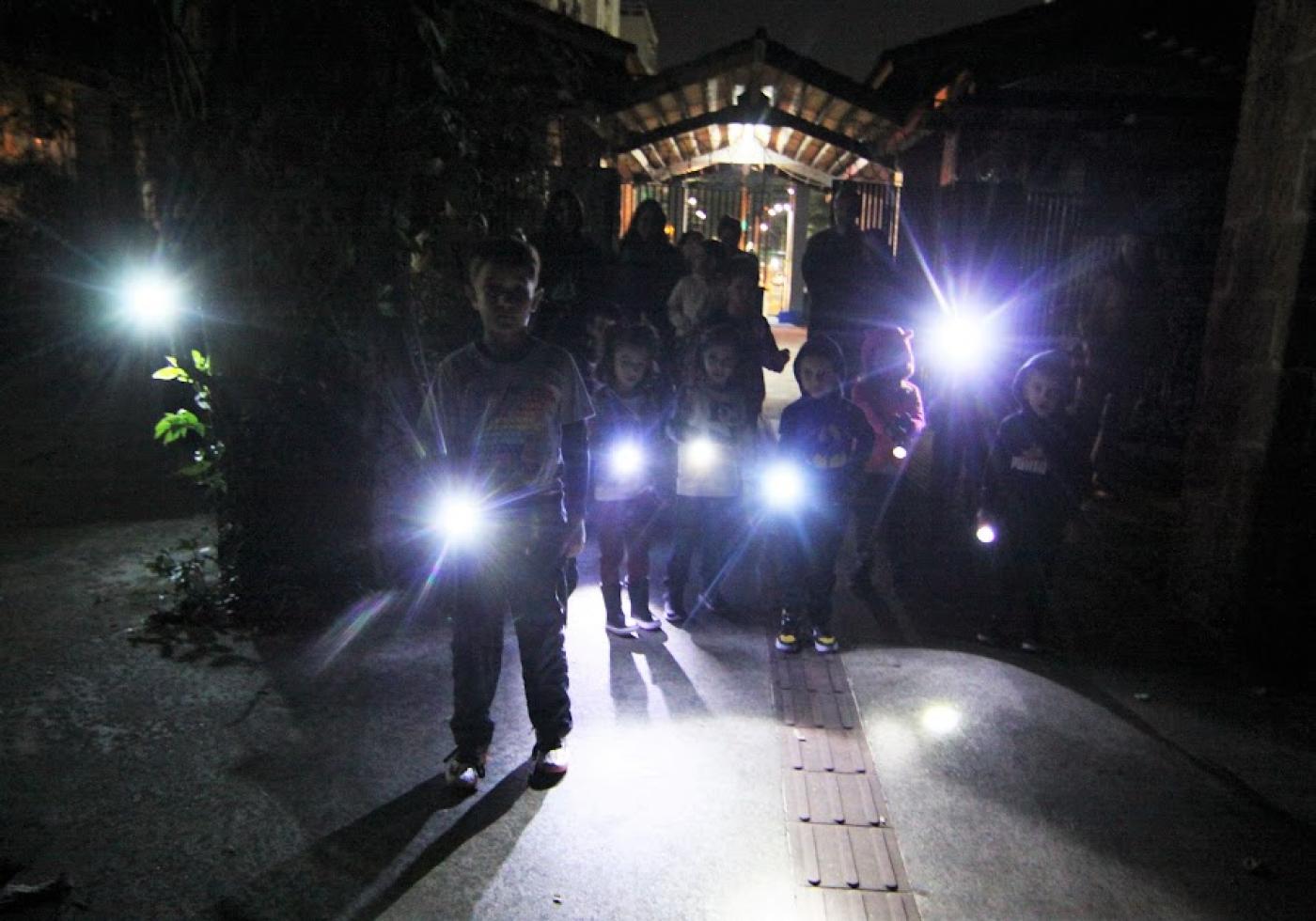 crianças seguram lanternas na entrada do orquidário. É noite. Ambiente está escuro. #paratodosverem 