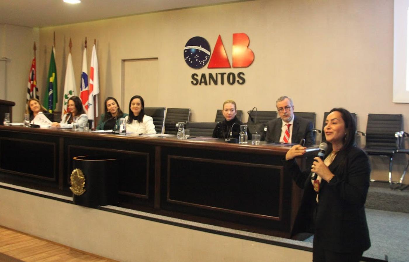 mulher fala ao microfone. Atrás dela ha um palco com seis pessoas sentadas diante de um balcão. Ao fundo, o símbolo da OAB. #paratodosverem 