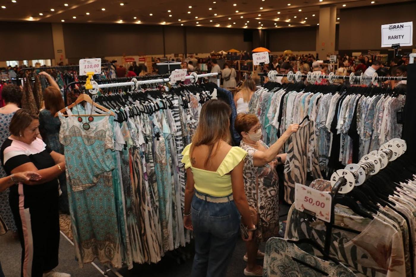 peças de roupas penduradas em araras e mulheres olhando e mexendo. Ambiente é de um grande bazar. #paratodosverem