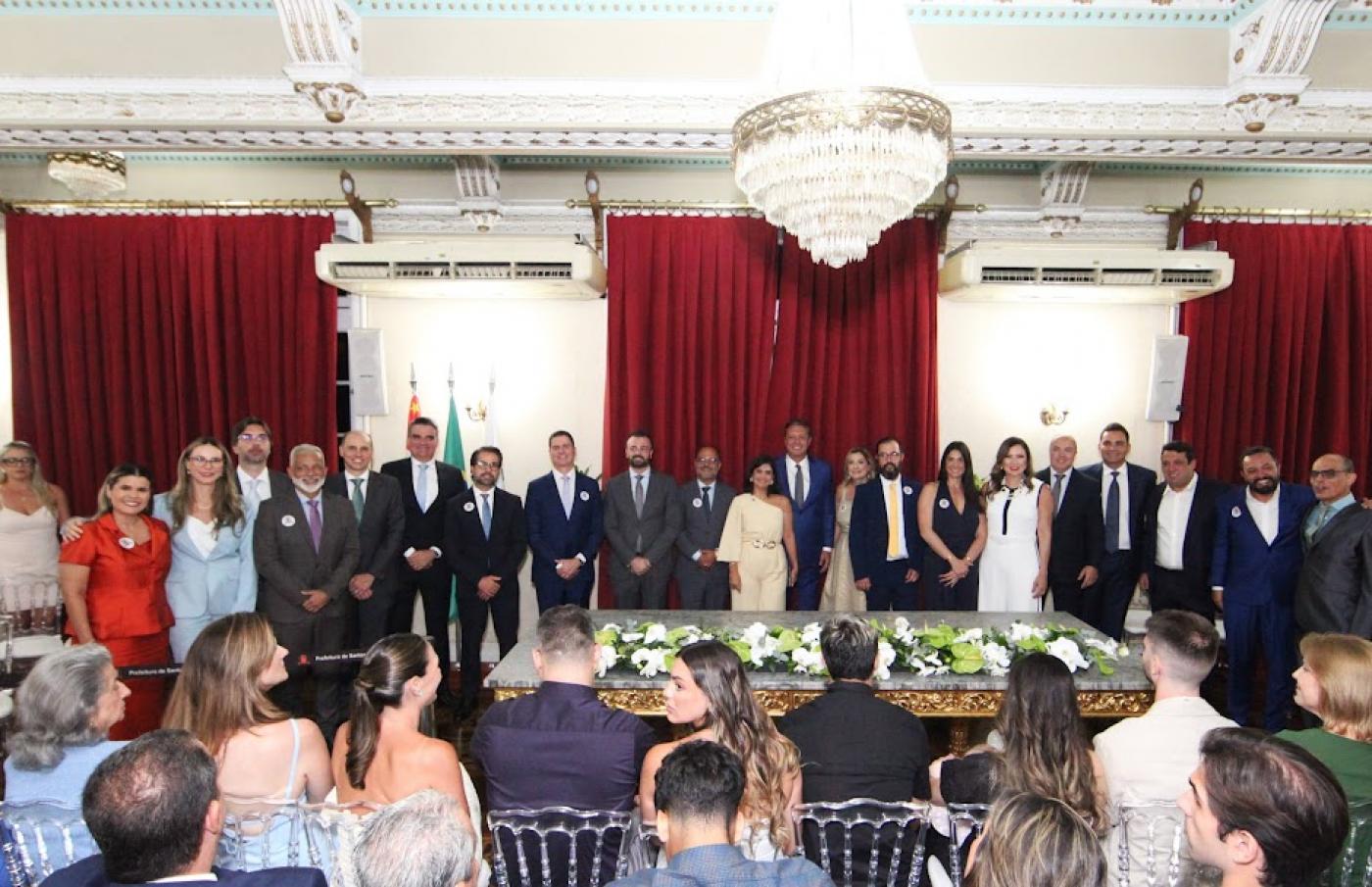 secretários, vice prefeita e prefeito em pé, posam para foto em auditório, com público sentado. #paratodosverem 