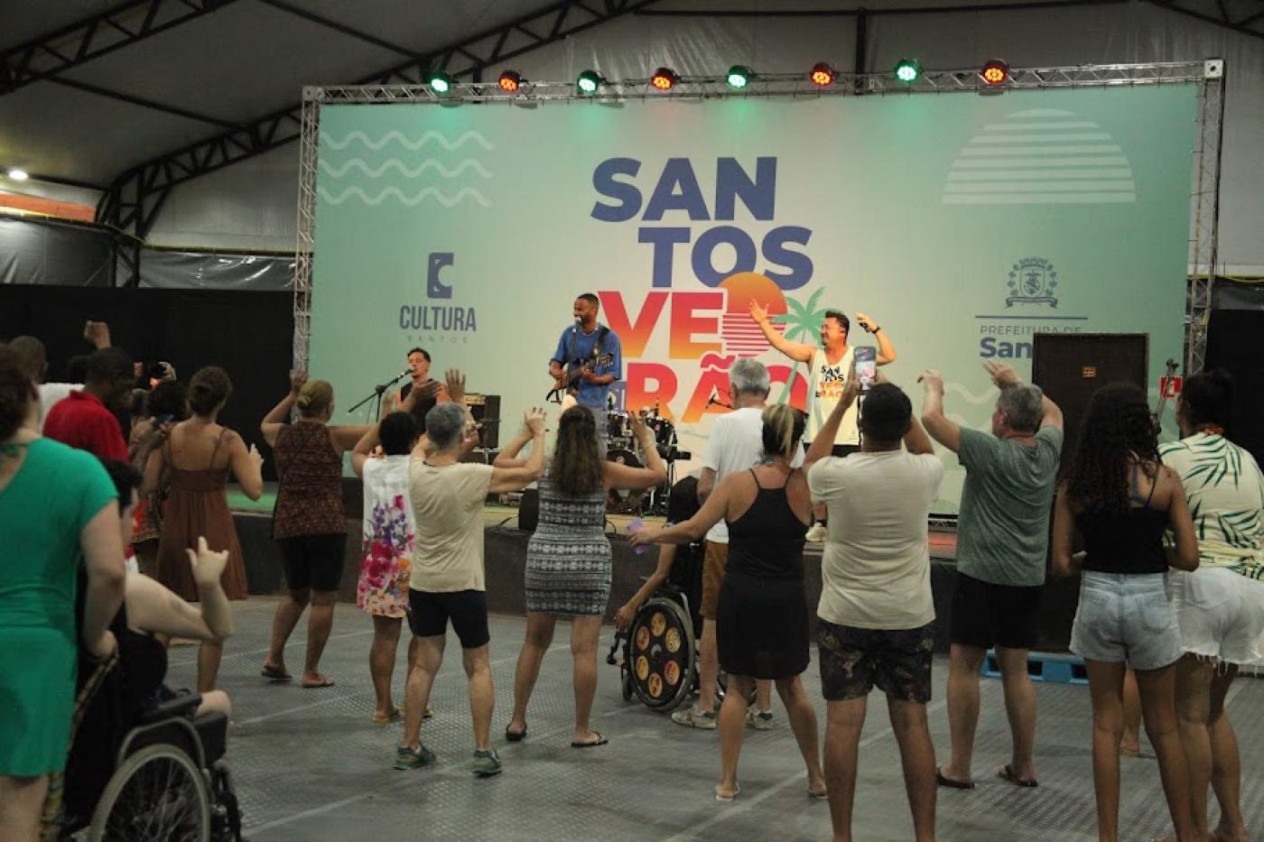 pessoas dança sob tenda, de costas para a foto e de frente para um palco onde um grupo se apresenta. #paratodosverem 
