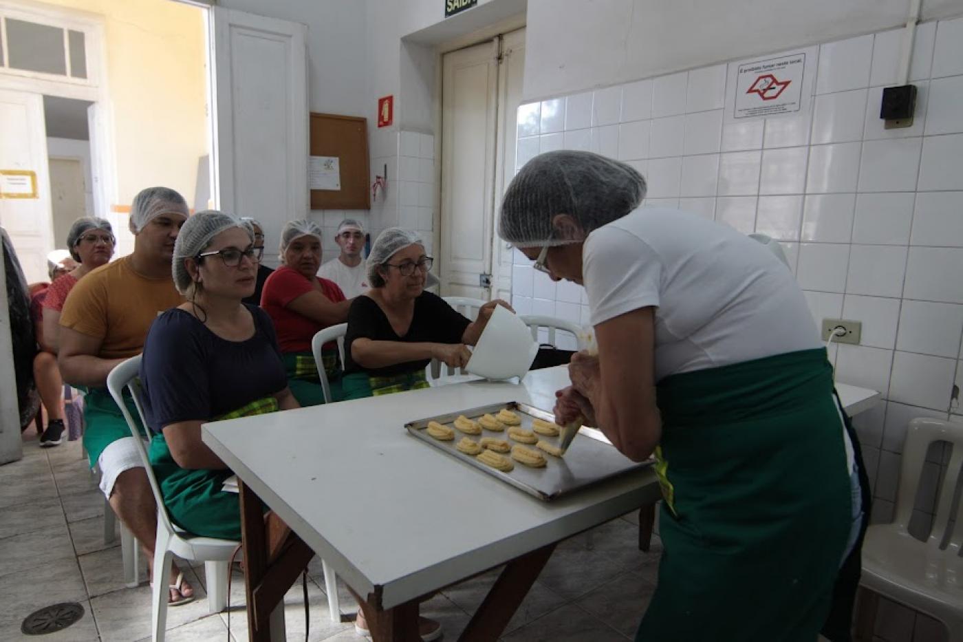 mulher está moldando pães sobre uma forma. Alunas ao fundo assistem. #paratodosverem
