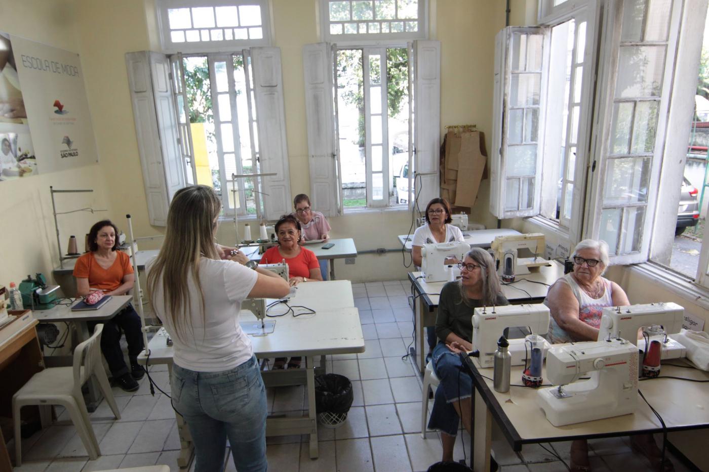 mulheres em sala de aula diante de máquinas de costura. #paratodosverem 