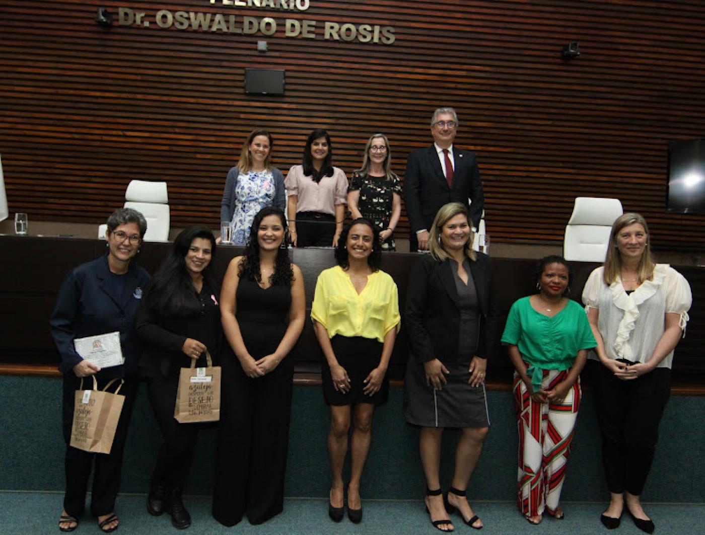 As sete homenageadas posam para foto. Todas em pé lado a lado. Ao fundo, no palco, autoridades municipais. #paratodosverem