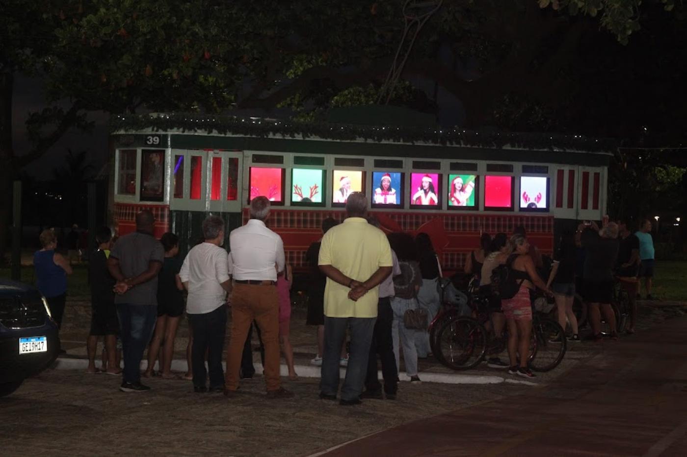 pessoas estão paradas diante do bonde que tem imagens sendo projetadas nas janelas. É noite. #paratodosverem 