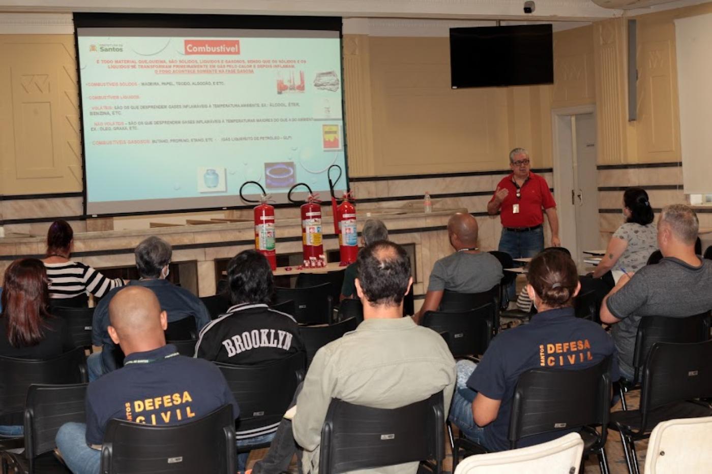 servidores estão sentados em auditório, de costas para foto, assistindo palestra. Há um telão na parede com textos sobre incêndio. Sobre uma mesa estão três extintores . #paratodosverem