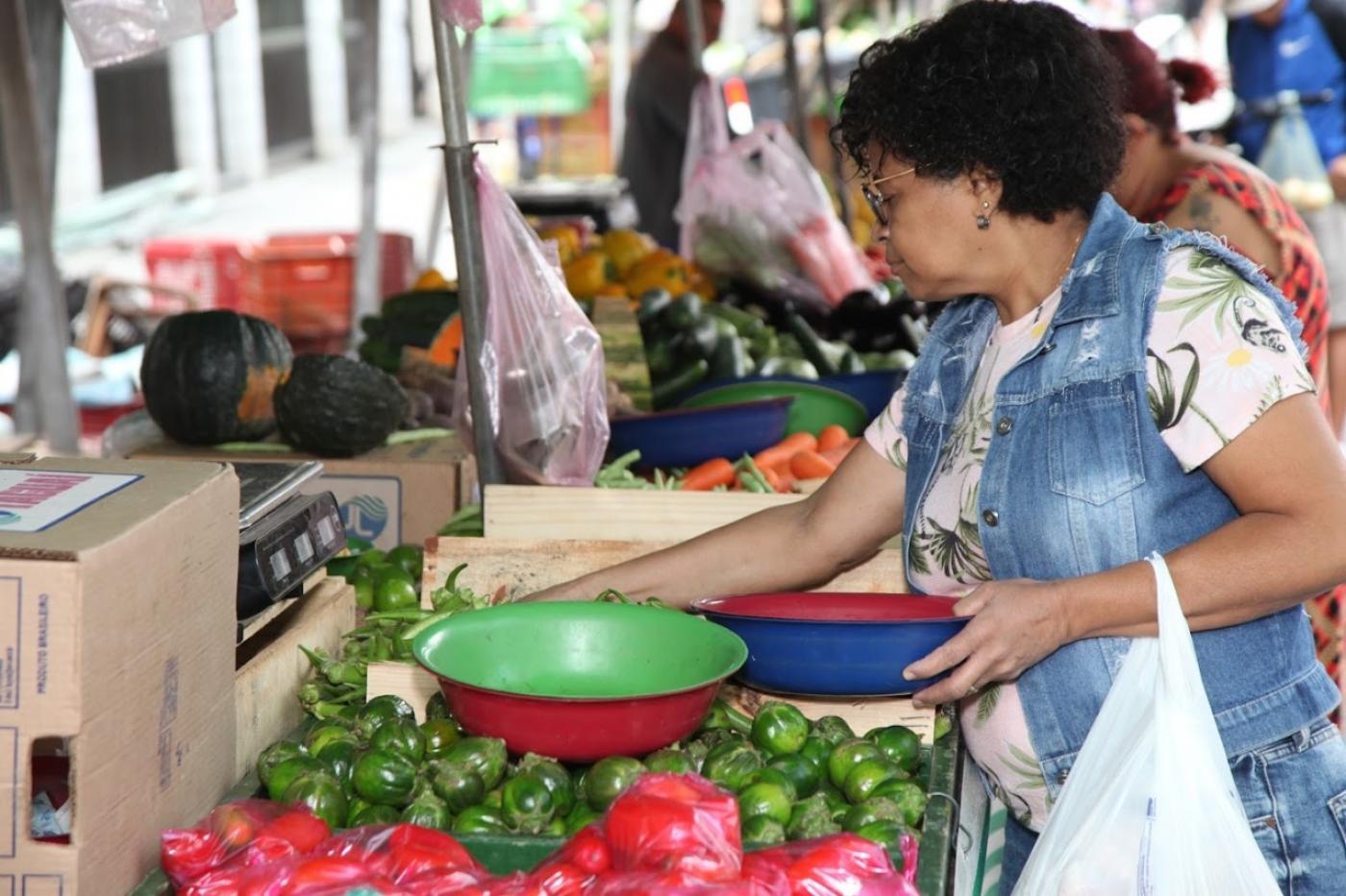 mulher está pegando limões em barraca de feira. Ela tem uma sacola pendurada no braço esquerdo. #paratodosverem