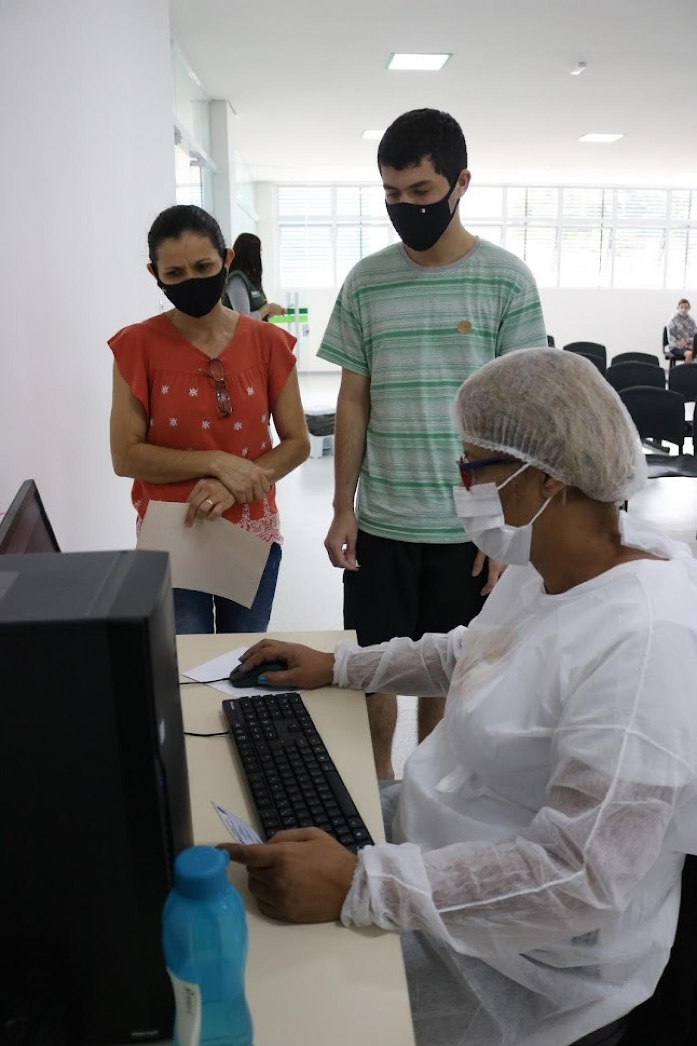 Pacientes em pé observam profissional de saúde operando computador. #pratodosverem