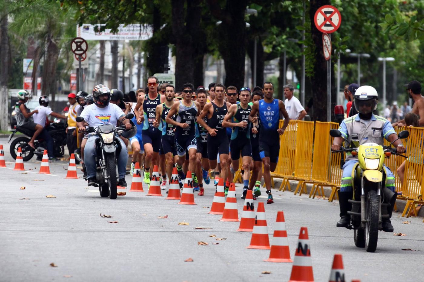 Atletas estão correndo em faixa de pista delimitada por cones. À frente e ao lado deles, dois motociclistas batedores. #Pracegover