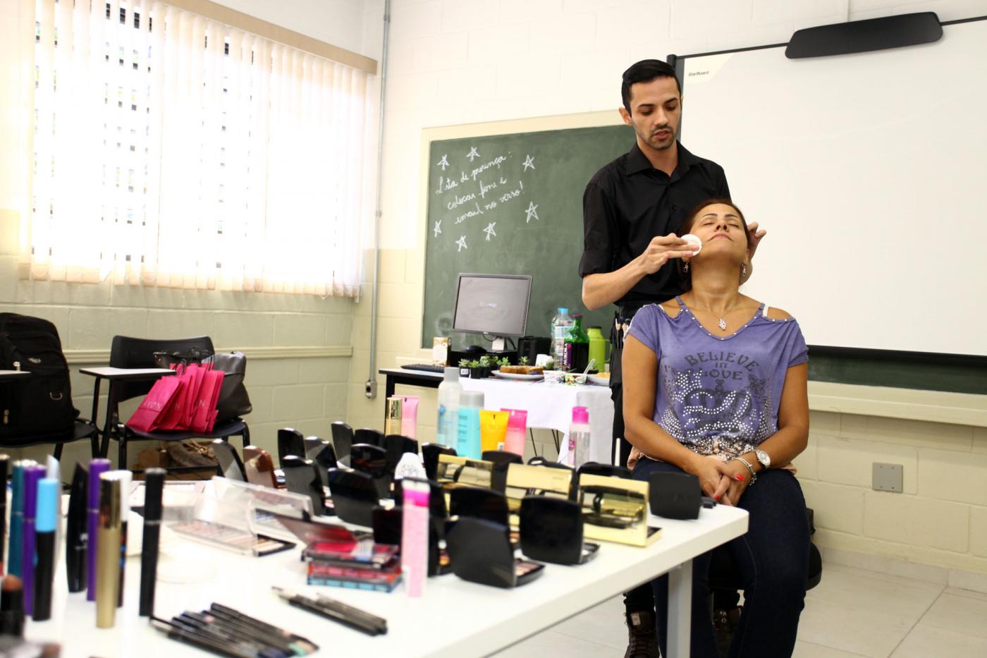 Mesa repleta de produtos de maquiagem. Um homem em pé está maquiando uma mulher que está sentada. #Pracegover mu