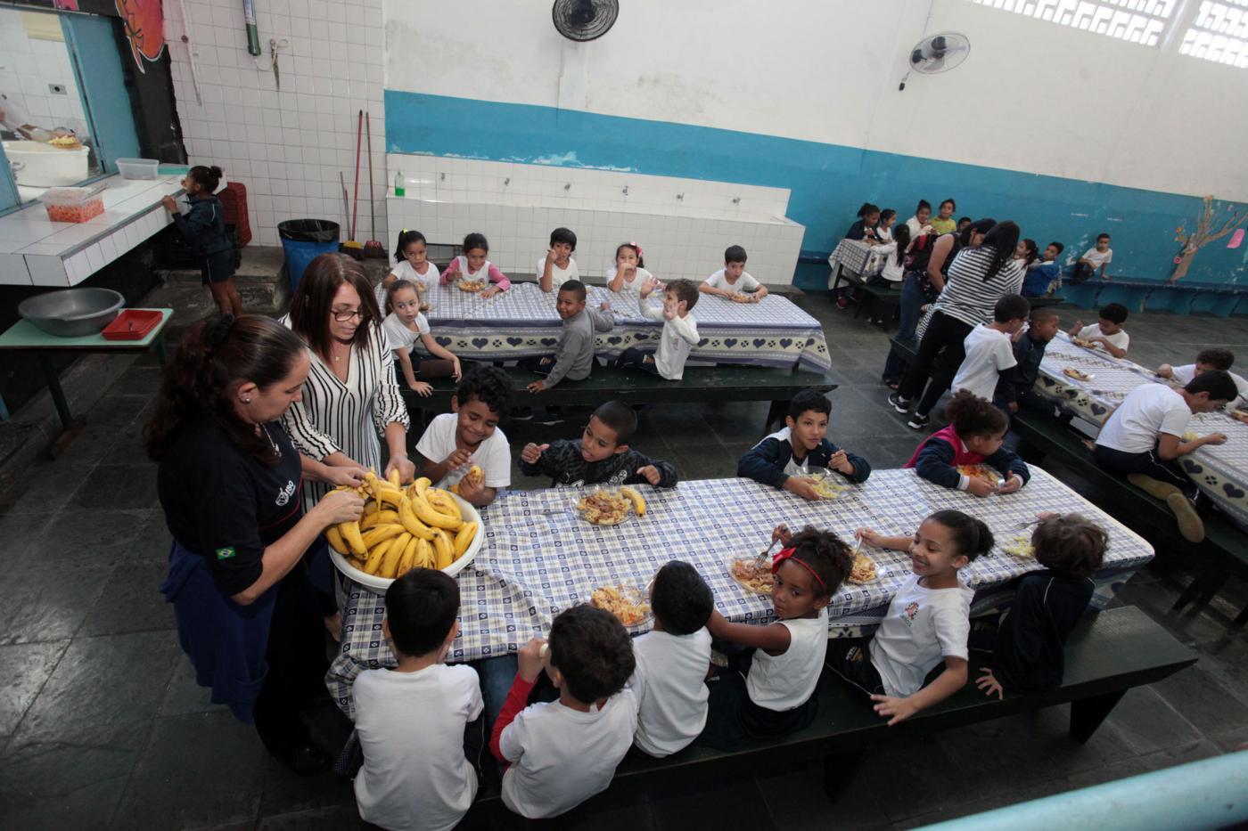 Alunos estão no refeitório tomando lanche. Todos estão sentados em torno de mesas. #Pracegover