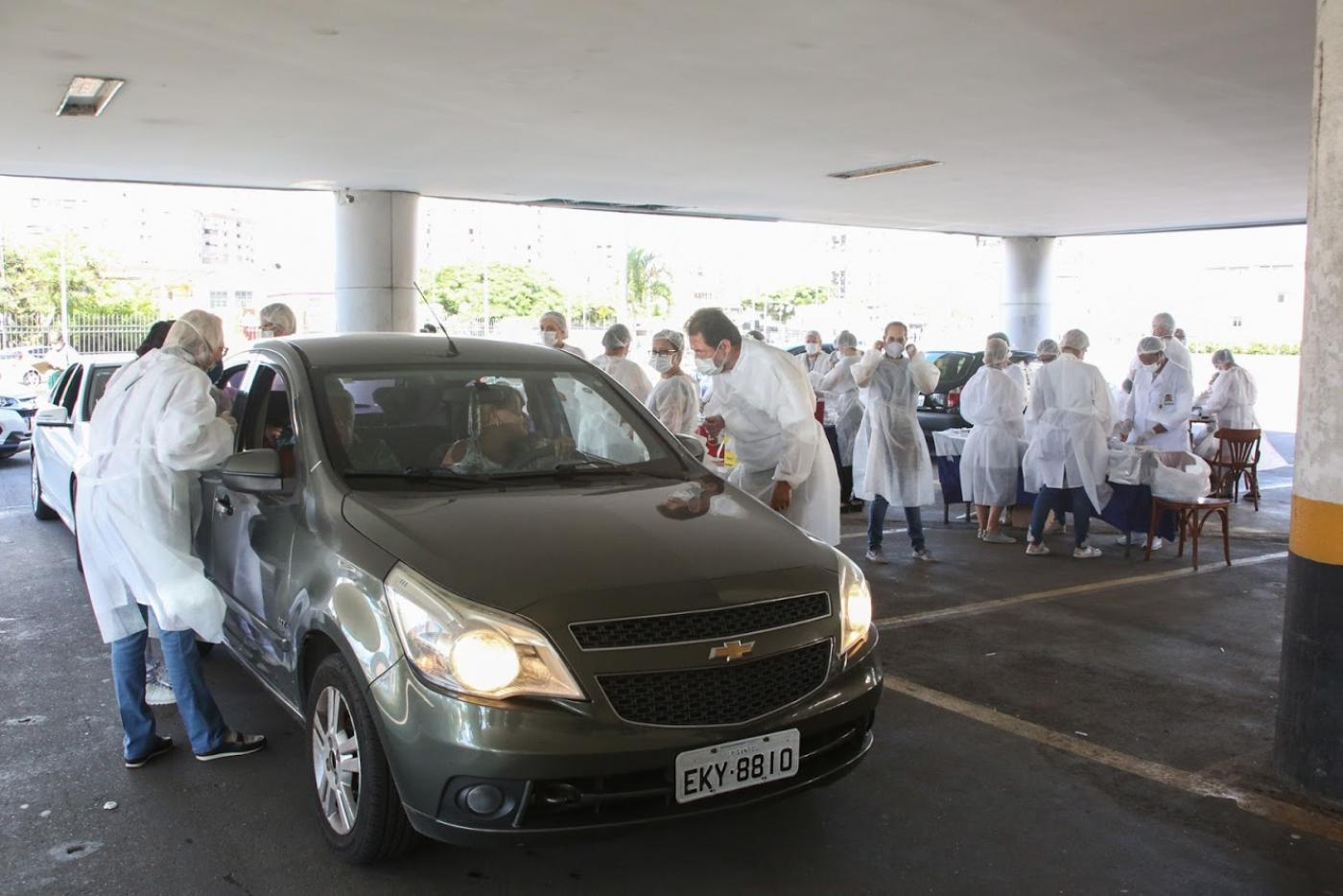 Veículo está parado em estacionamento. Pessoas vestidas aventais brancos, máscaras e luvas estão em volta. #Paratodosverem