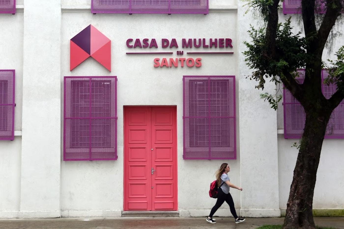 fachada da casa da mulher com uma mulher passando à frente, na calçada. Há também o troco de uma grande árvore. #paratodosverem 