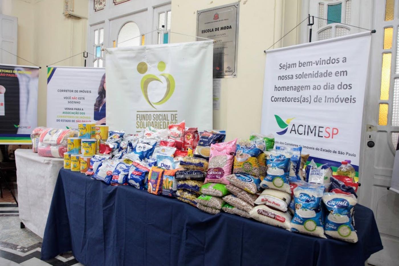 grande volume de alimentos não perecíveis sobre uma mesa com banner da acimesp e do fundo social ao fundo. #paratodosverem