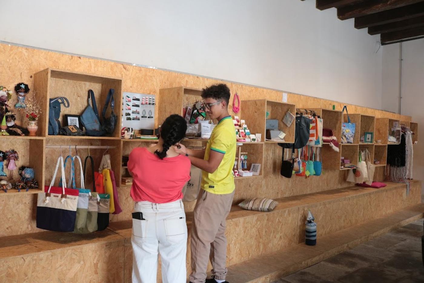 homem e mulher estão de costas para a foto e de frente para prateleira onde estão expostos produtos artesanais. #paratodosverem 