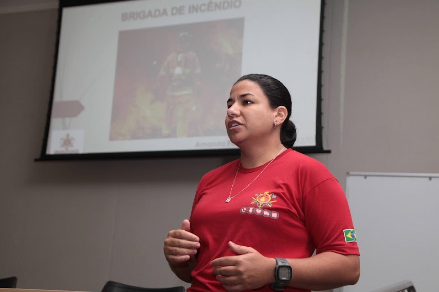 bombeira está falando diante um telão com imagem de incêndio projetada. #paratodosverem