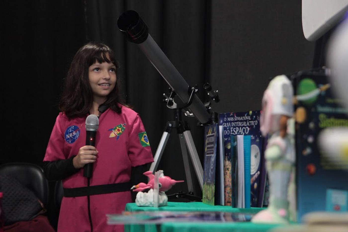 menina está segurando microfone ao lado de mesa com telecópio e outros objetos que tem ligação com o espaço. #paratodosverem