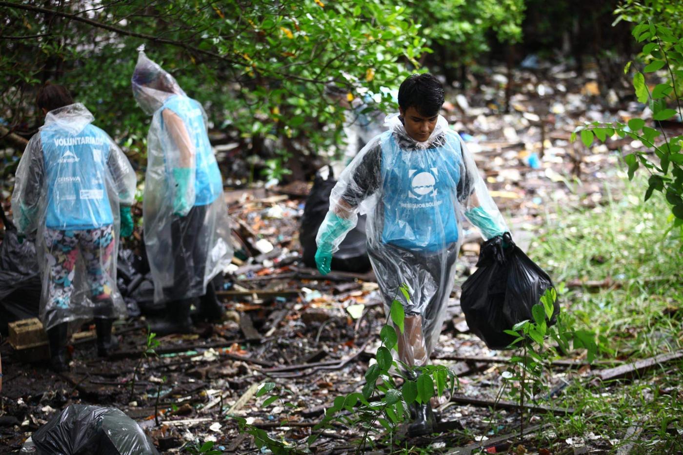 JOvens usando luvas estão em mangue recolhendo residuos e depositando em sacos. #paratodosverem