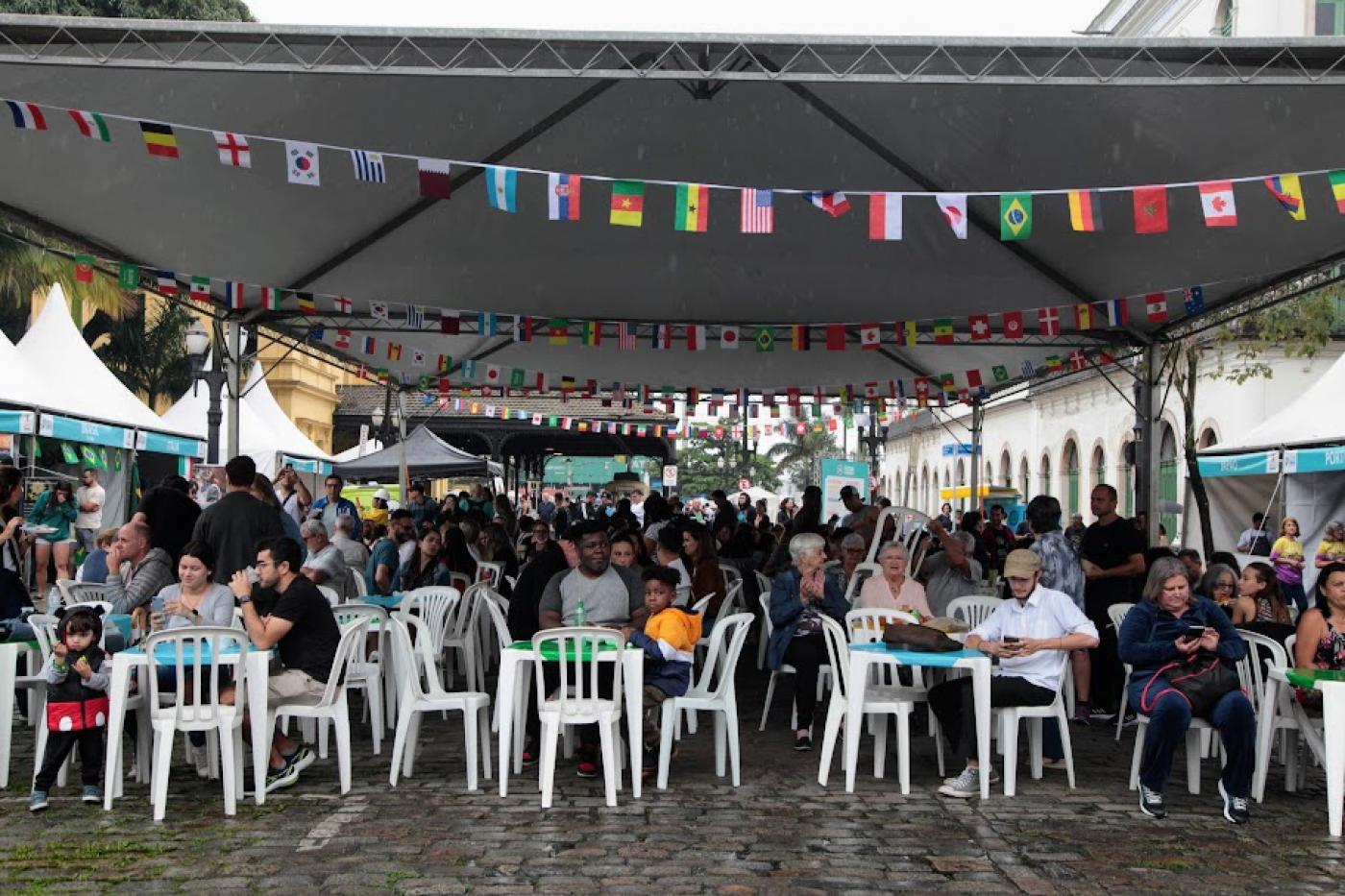 pessoas estão sentadas em praça de alimentação decorada com bandeirinhas de diversos países. #paratodosverem 