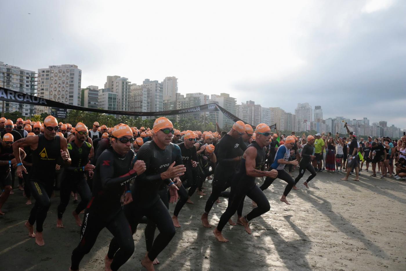atletas uniformizados com roupa de natação e toca correm na praia em direção ao mar para prova de nado. #paratodosverem 