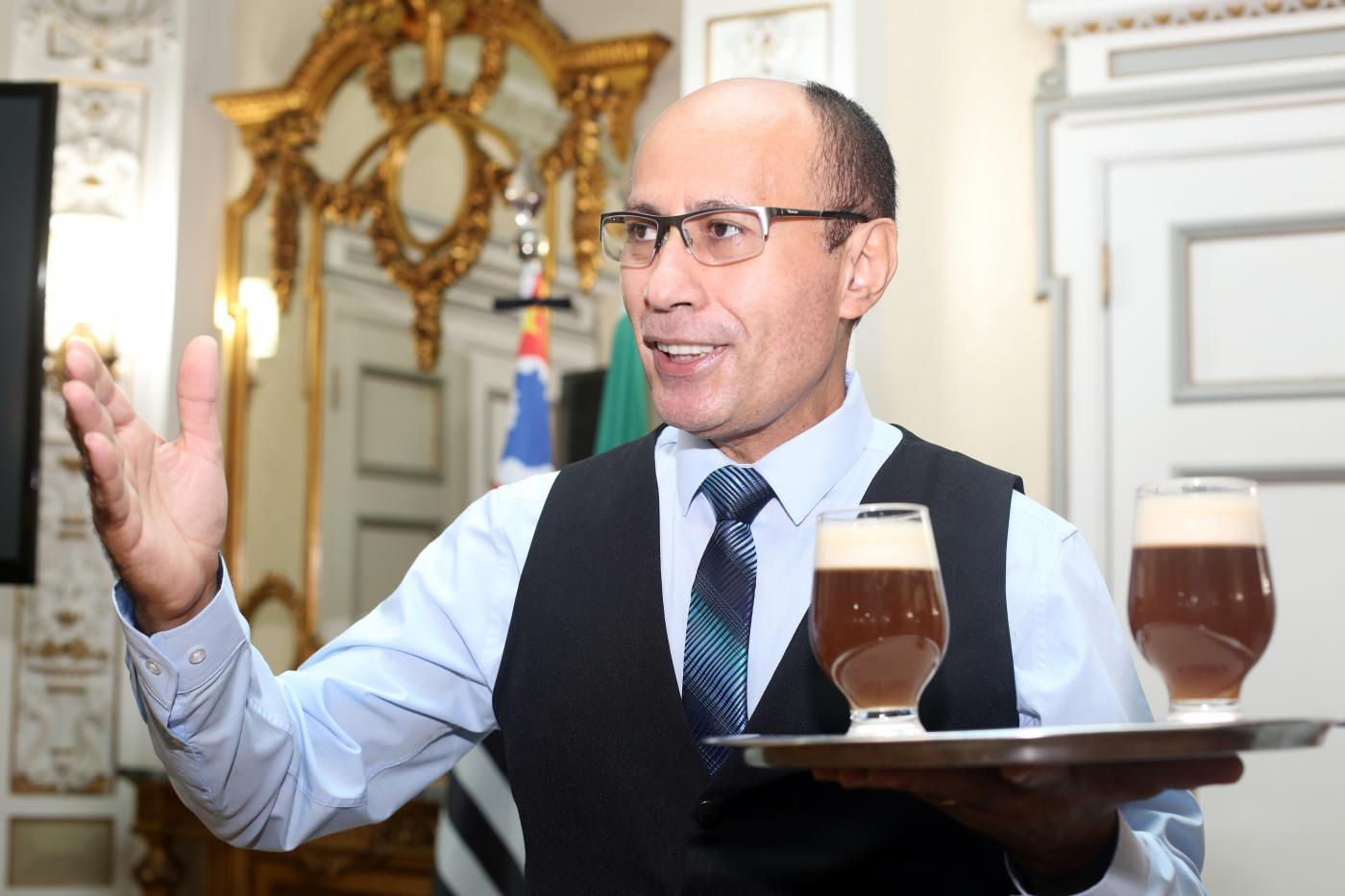 #pracegover no salão nobre da prefeitura, João segura bandeja com dois copos de chá