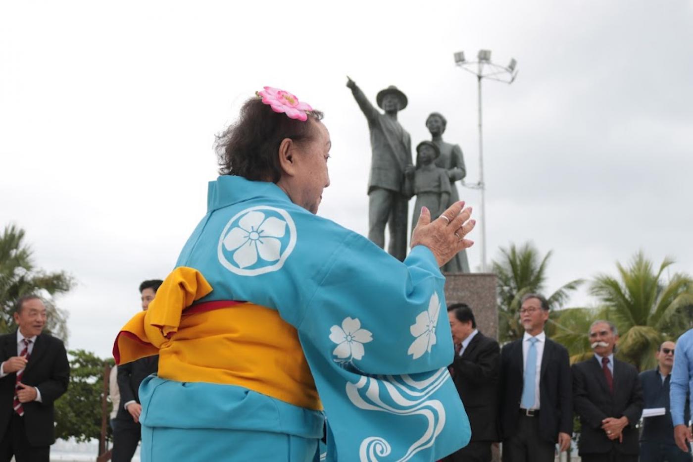 Mulher vestida com roupa típica dança em frente a monumento em homenagem à imigração, com estátuas de três pessoas formando uma família. #paratodosverem