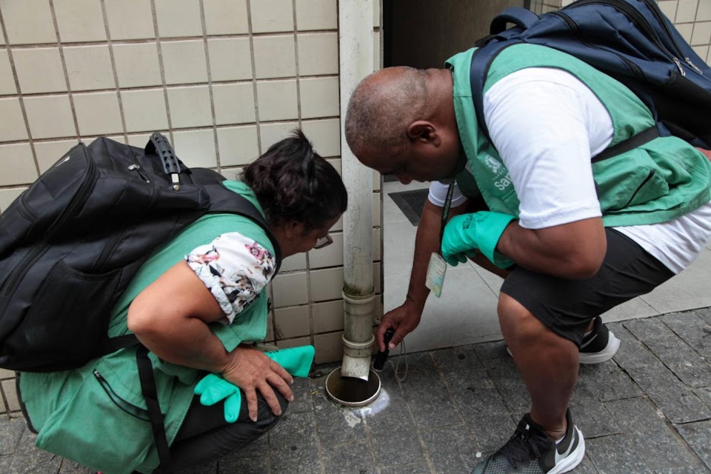 dois agentes observam saída de cano de água para ralo. #paratodosverem