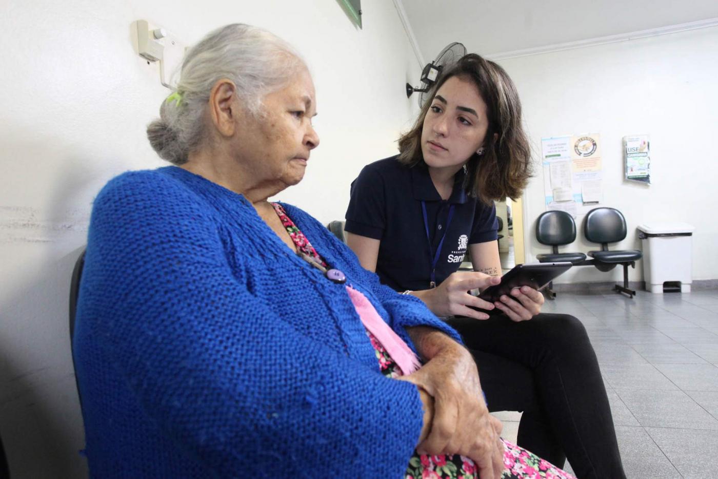 Estagiária da Ouvidoria conversa com idosa usuária de policlínica. Ambas estão sentadas. A jovem faz anotação em um tablet