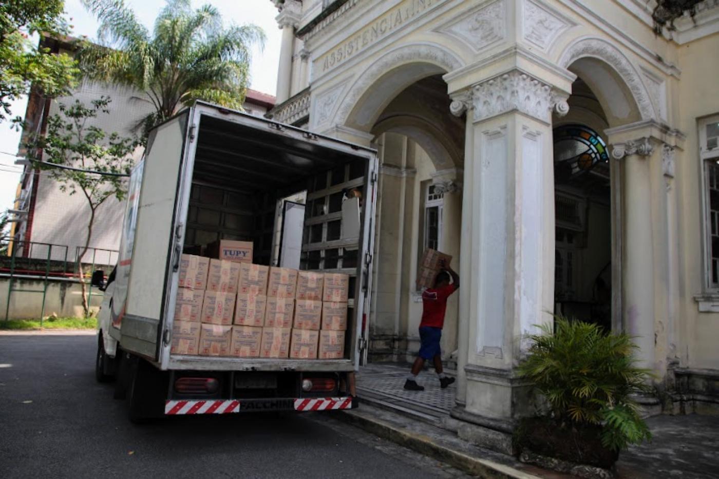 caminhão cheio de cestas básicas com a porta de trás aberta. Ao fundo, um homem carrega uma caixa para dentro de um prédio. #paratodosverem