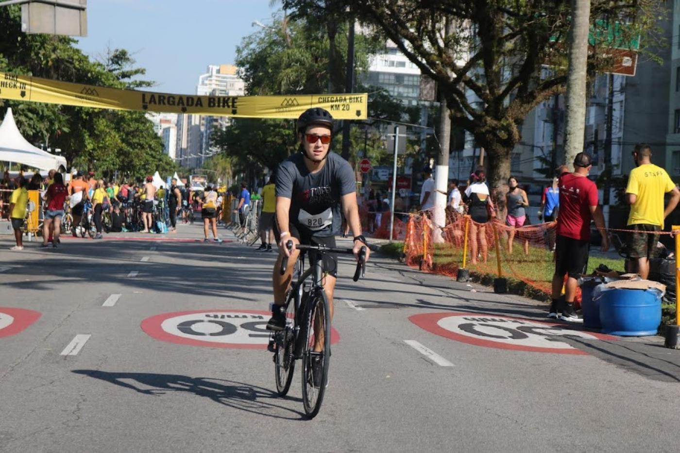 ciclista está pedalando em avenida da orla. Ao fundo há várias pessoas. #paratodosverem