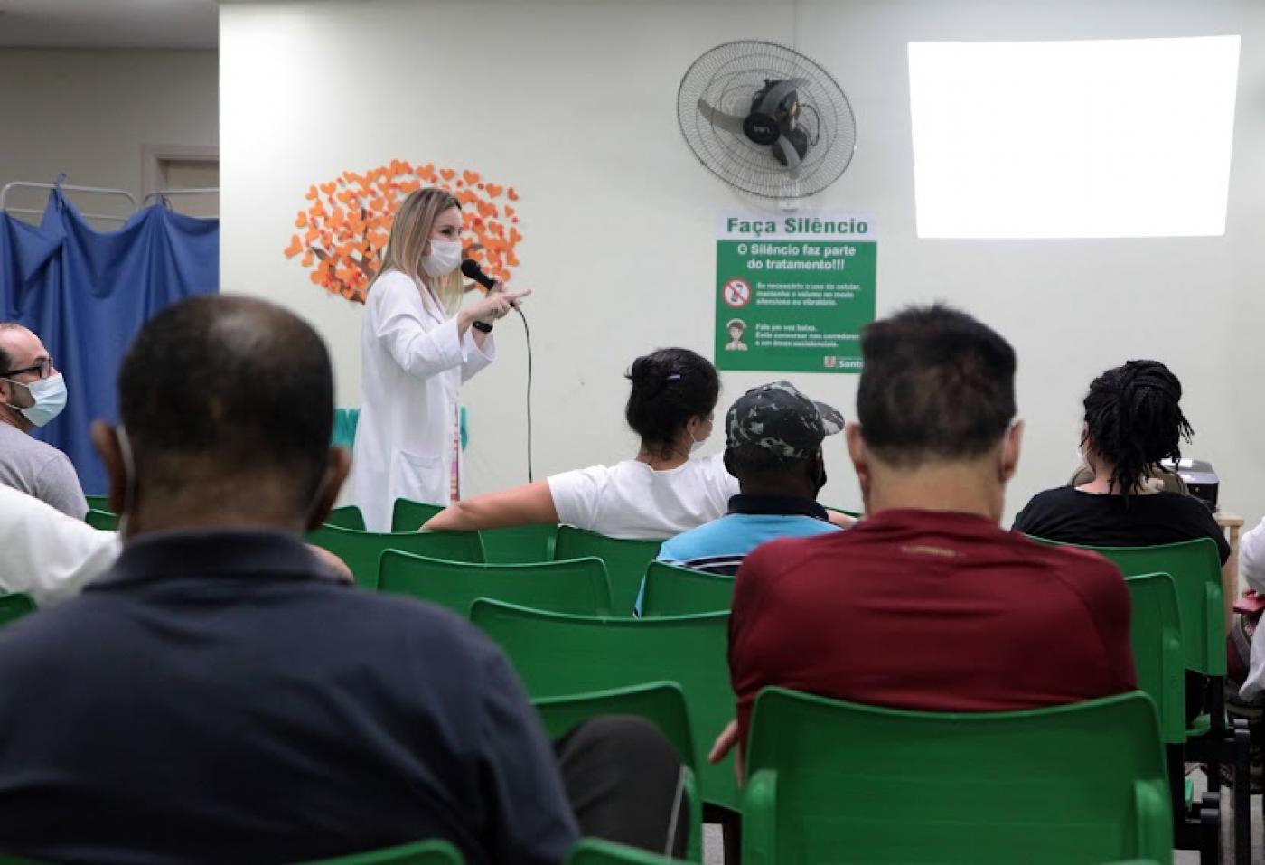 pessoas estão sentadas em sala de espera assistindo palestra feita por uma mulher. #paratodosverem