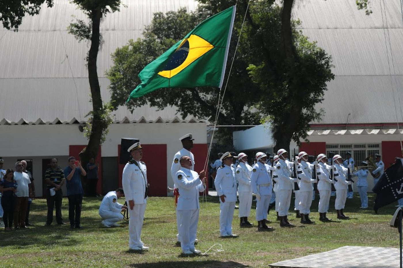 marinheiros uniformizados hasteando a bandeira do brasil ao ar livre sobre um gramado. #paratodosverem 