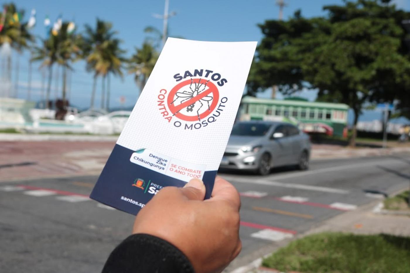 close sobre mão direita que segura folheto onde se lê: Santos contra o mosquito. A avenida da praia está ao fundo. #paratodosverem 