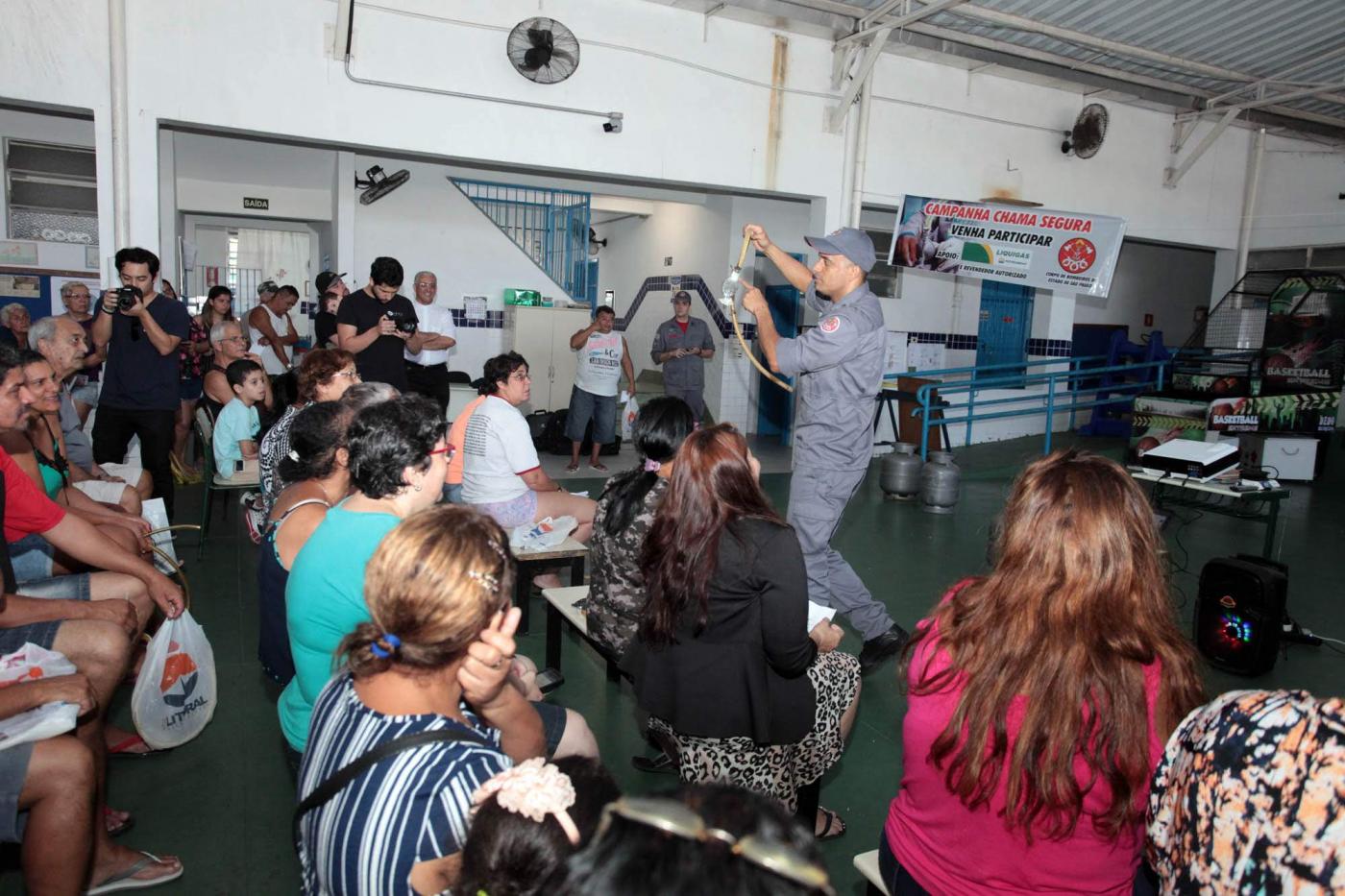 Cerca de 20 pessoas sentadas observam bombeiro uniformizado que mostra mangueira e válvula de botijão de gás