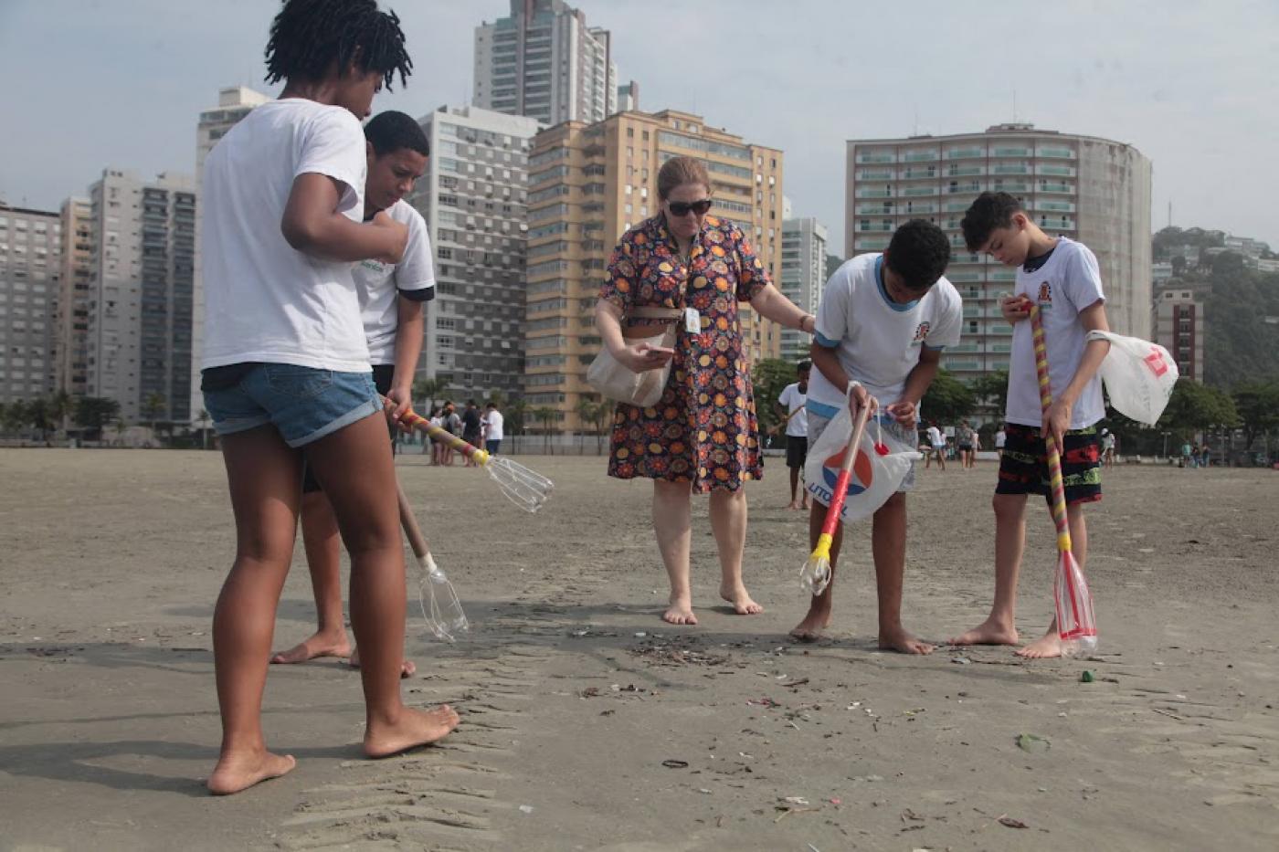 estudantes uniformizados andam na areia da praia catando lixo com uma garra feita com tubo pvc e garrafa pet. #paratodosverem 
