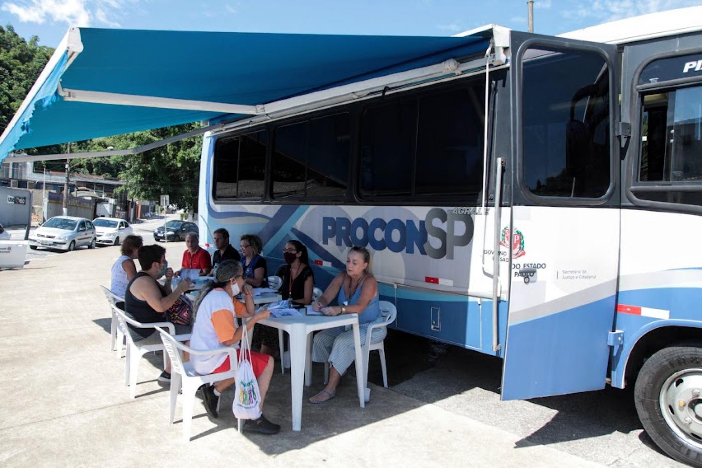 Ônibus do procon móvel está parado. Pessoas estão sentadas sendo atendidas por recepcionistas. #paratodosverem