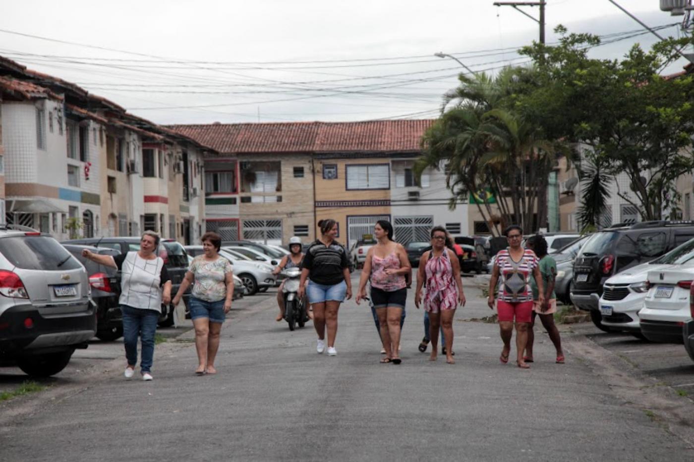 mulheres caminham por rua de uma vila. Nas laterais, os carros estão estacionados a 90 graus. #paratodosverem 