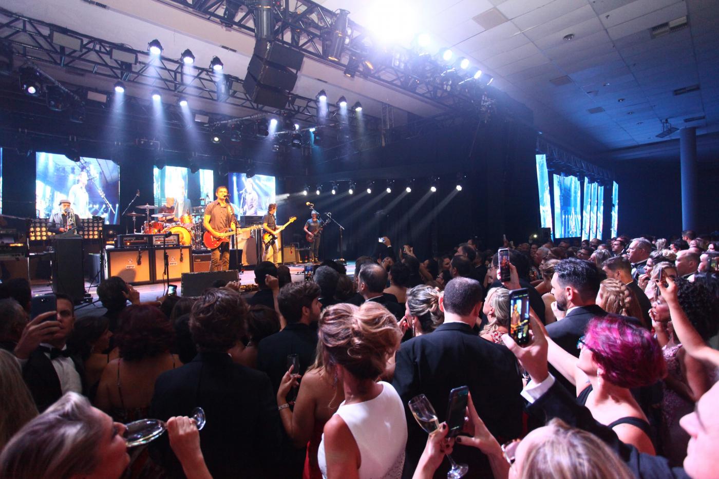 Baile com pessoas dançando em pista. À frente tem um palco com um cantor. #Pracegover