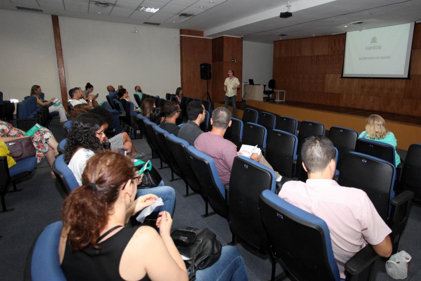 Pessoas estão sentadas em poltronas de auditório. Elas estão atentas a um homem que fala ao fundo. Na parede há uma imagem projetada em um telão. #Pracegover