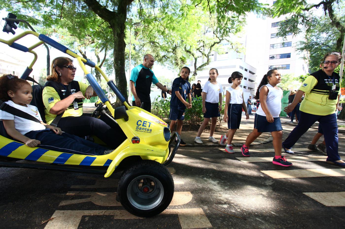 Minibugue aparece em primeiro plano com uma mulher pilotando e uma criança sentada ao lado. À direita, crianças atravessam na faixa. #Pracegover