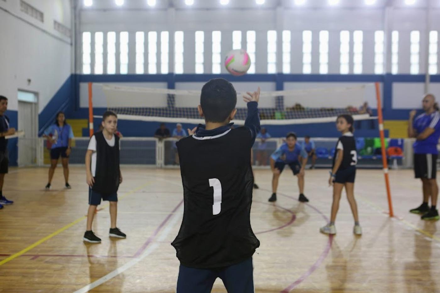menino de costas para a foto está no início da quadra lançando a bola para o outro lado da rede. #paratodosverem 