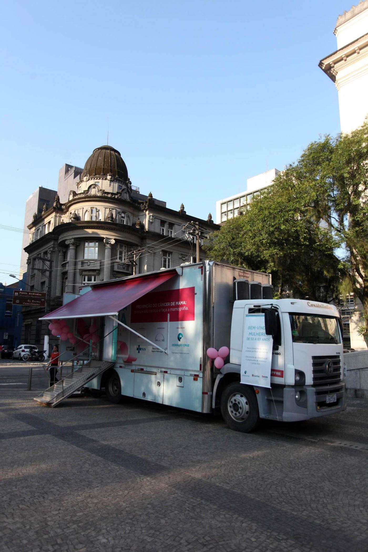 Caminhão estacionado em praça. O veículo tem  uma tenda na frente e uma rampa de acesso ao seu interior. #Pracegover