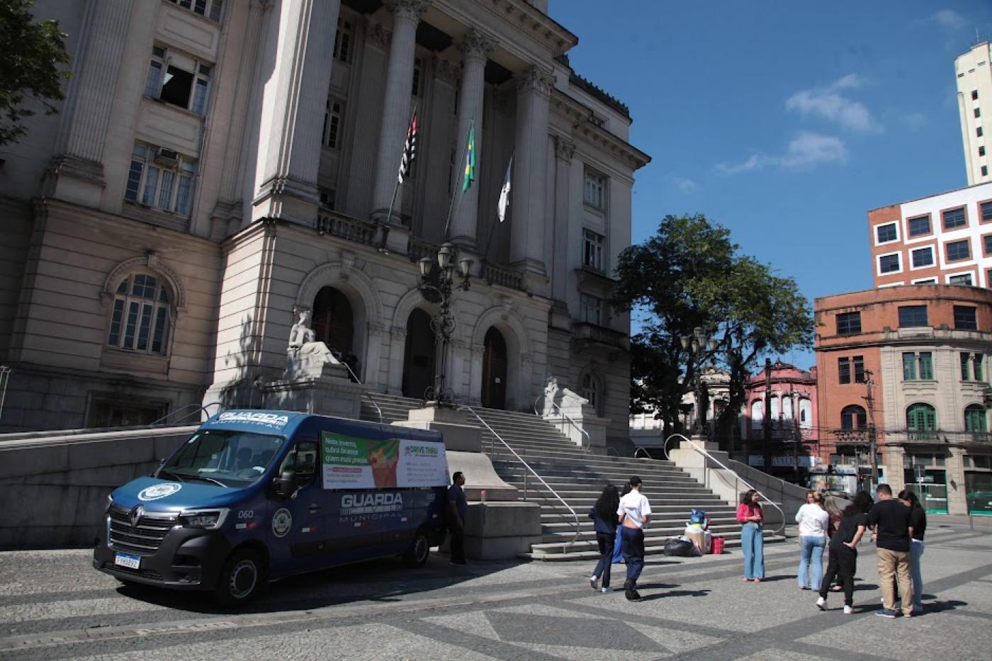 van da guarda municipal está parada na praça mauá em frente ao Paço. Ao lado, vários pacotes de roupas e pessoas circulando. #paratodosverem