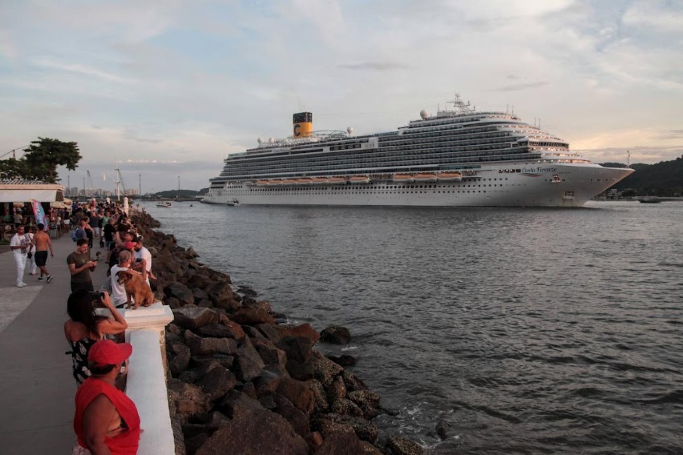 navio está saindo do porto de santos e pedestres estão próximos à mureta da ponta da praia para avistá-lo. #paratodosverem 