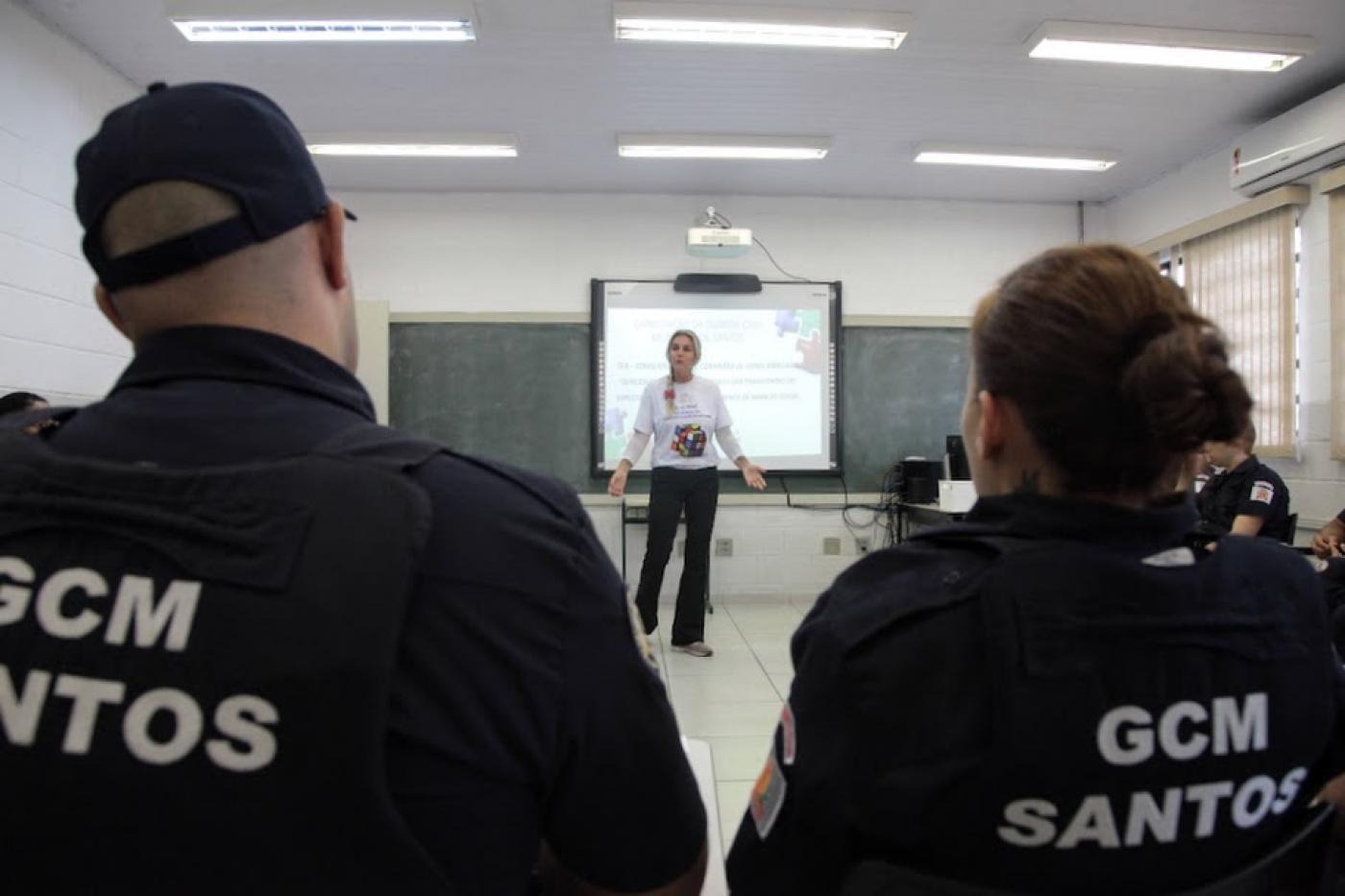 dois guardas municipais com coletes, sentados, de costas para a foto. Ao fundo, aparecendo entre o meio dos ombros deles dois, há uma mulher falando à frente de um telão. #paratodosverm