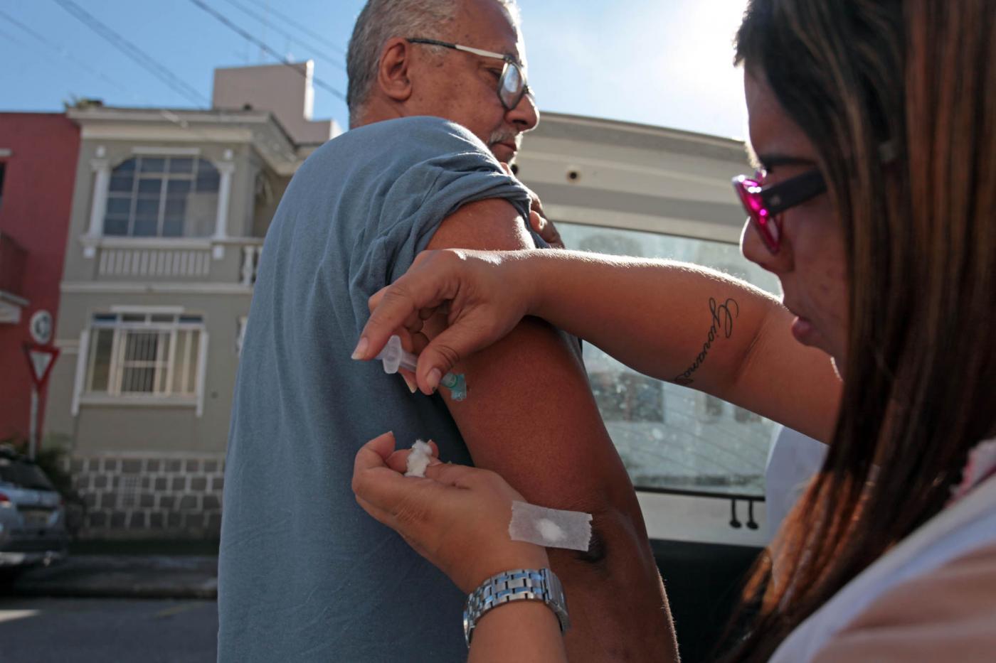 Agente da saúde vacina homem. Ao fundo se vê alguns sobrados na rua. #Pracegover