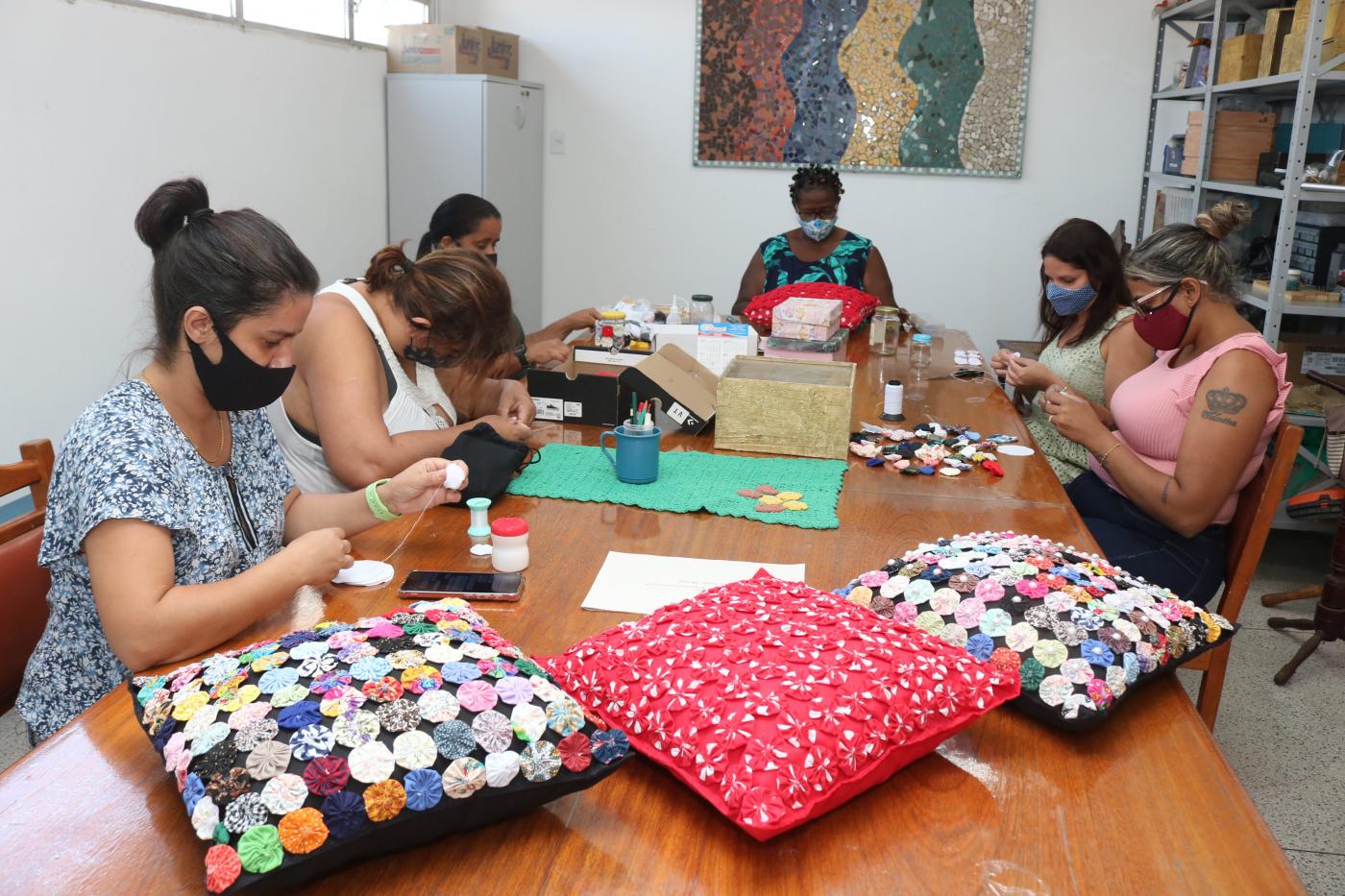 seis mulheres sentadas costurando sobre uma mesa , almofadas sobre a mesa #paratodosverem