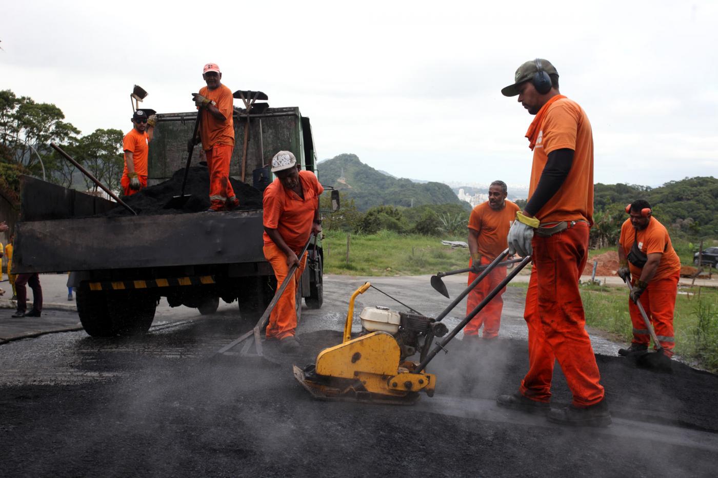 Homens uniformizados aplicam massa e nivelam solo com pás e máquina. Há um caminhão atrás dos trabalhadores. #Pracegover