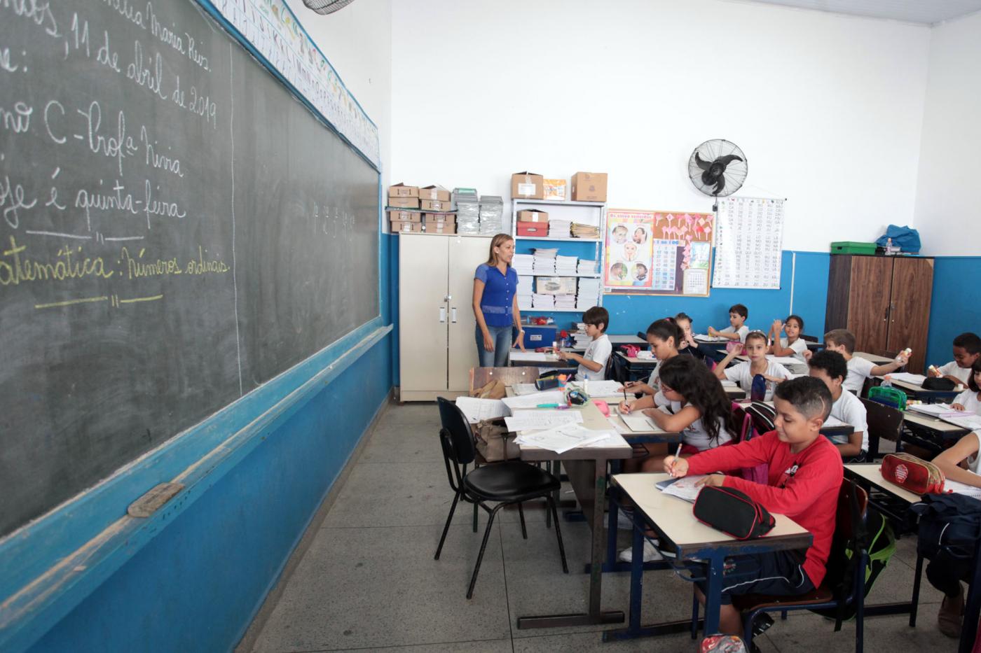 Sala de aula com professora ao fundo. A lousa está ao lado esquerdo da imagem. Os alunos estão sentados em suas carteiras. #Pracegover