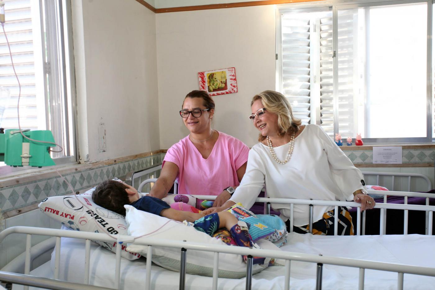 Criança está em maca de hospital. Duas mulheres estão ao seu lado e fazem carinho na criança. #Pracegover